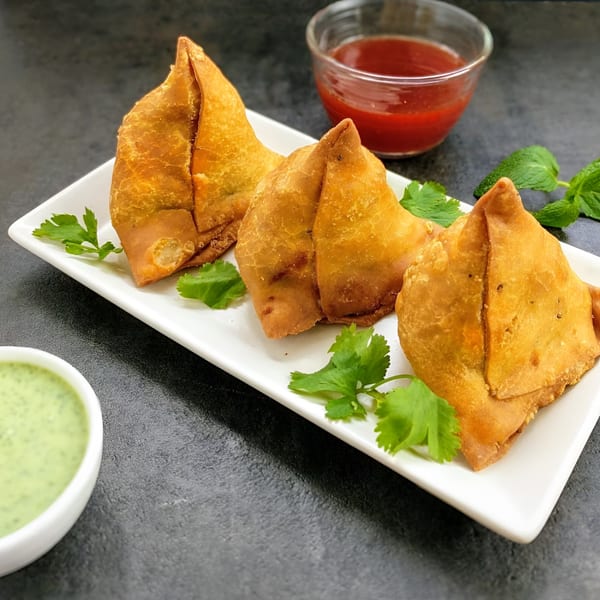 Three golden-fried samosas garnished with cilantro and mint, served with red and green chutneys.