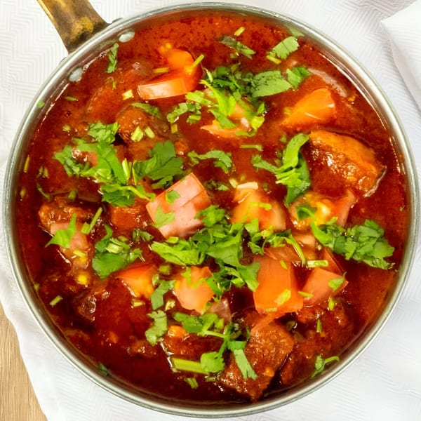 Dhabewala Indian Shack's chicken curry, garnished with fresh cilantro and diced tomatoes, in a metal pot.